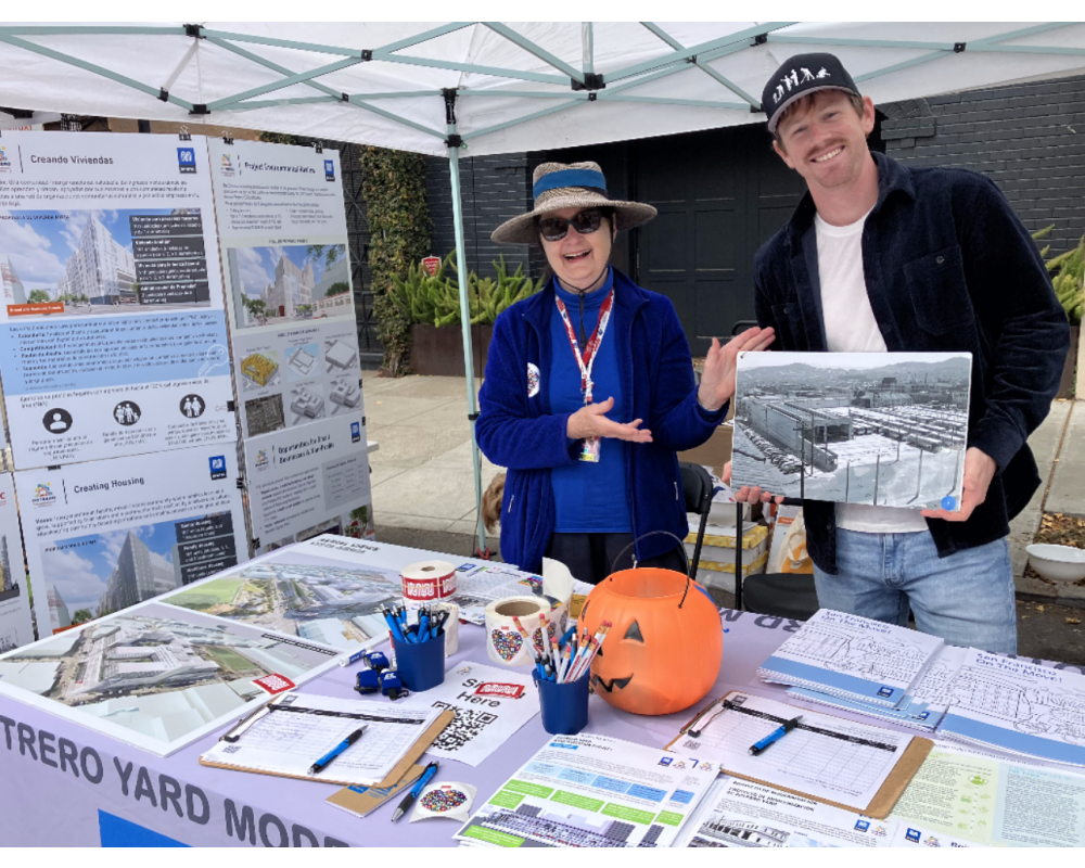 Two people looking at the camera while smiling and gesturing towards visual and printed information on a table
