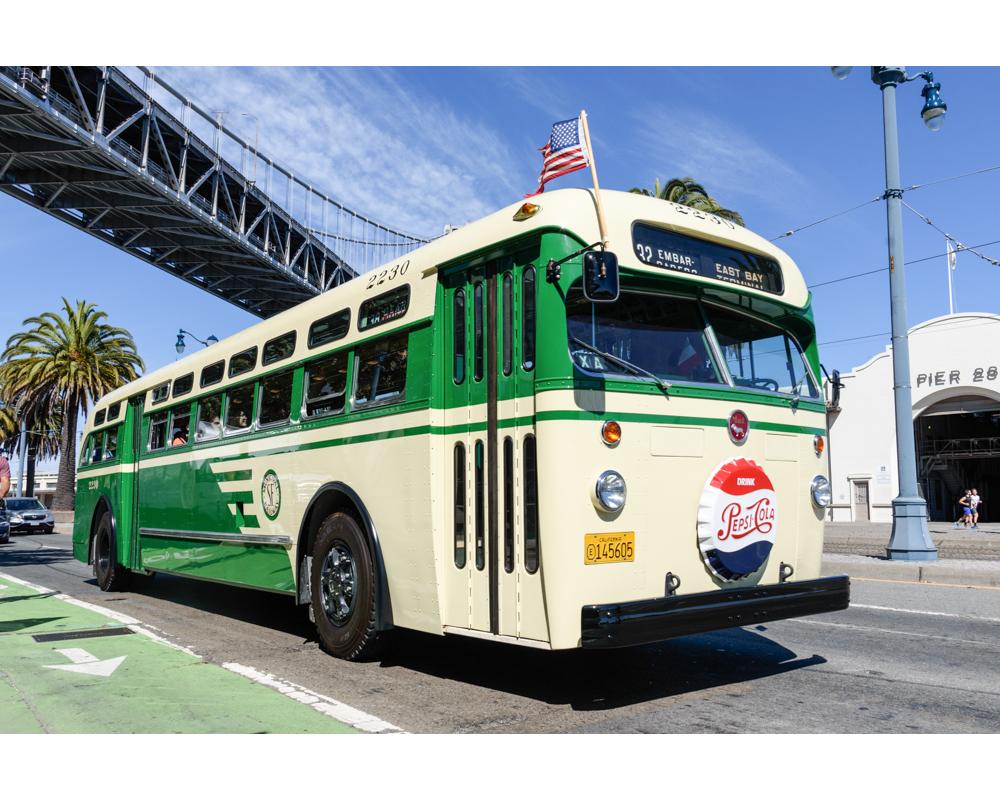 A vintage Muni bus that's green and cream in color passing under a bridge 