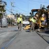 Crew member by the rail track and an excavator with a shovel along the street with houses lined along the side. 