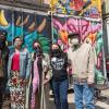 Several individuals with masks huddle for a group photo in front of a colorful mural backdrop in an our setting. 