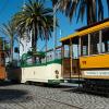 An orange historic streetcar passes a green and cream open-air Boat Tram and another orange historic streetcar. Palm trees and Ferry Building in the background.