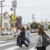 Several people cross a street near a yellow rapid flashing beacon. It includes a yellow sign and one with an arrow.