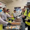 people in safety vests are in line in front of large dishes of food  Smiling people are serving them.