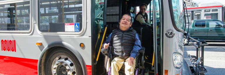 A customer in a wheelchair exits a bus while a driver deploys the lift