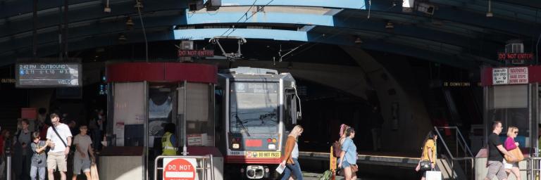 Muni train approaches exit of West Portal Station as people walk in the crosswalk in front of the train.