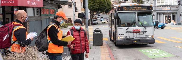 Two SFMTA staff working with a San Francisco resident on a survey with a bus approaching