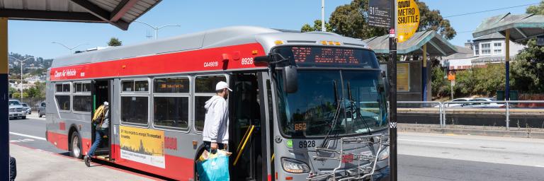 A 28R 19th Avenue Rapid bus boards outside of SF State University. Two people walk on the bus at the front and back doors.