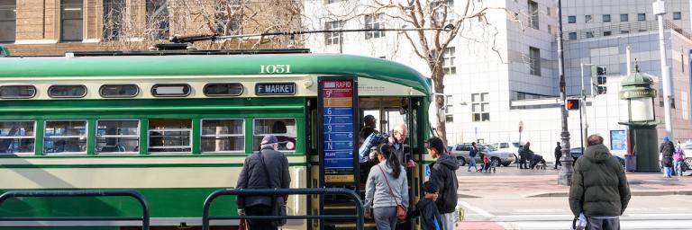 A green and cream F Market streetcar stops at 8th and Market across the street from the Main Branch Library. People wait to board the streetcar.
