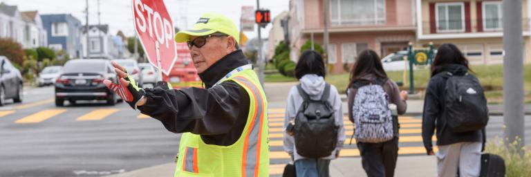 Crossing guard wearing yellow vests helps three students cross the street.