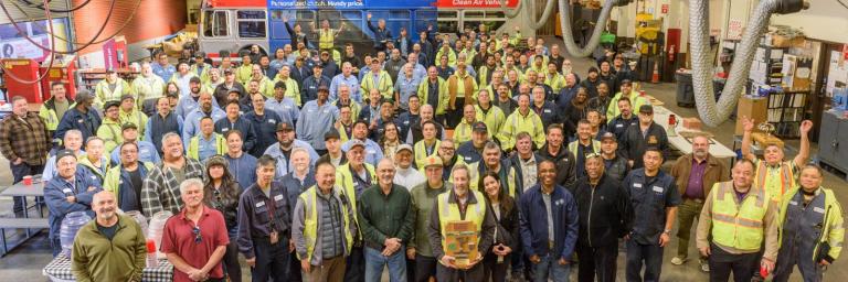 Large group of SFMTA staff in a bus yard. Many wear yellow vests.