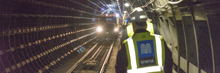 Two people in safety vest walking down subway tunnel with service truck in the distance
