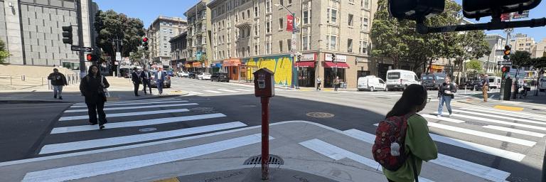 Pedestrians using new crosswalk at Larkin and Golden Gate intersection. One person waits near the traffic signal near the yellow tactile domes in the curb ramp leading to the crosswalk.