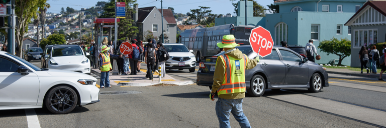 SFMTA Crossing Guard assisting pedestrian safety at intersection with cars and a Muni Metro Stop