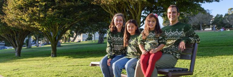 Family sitting on a park bench wearing the 2026 Muni Holiday Sweater