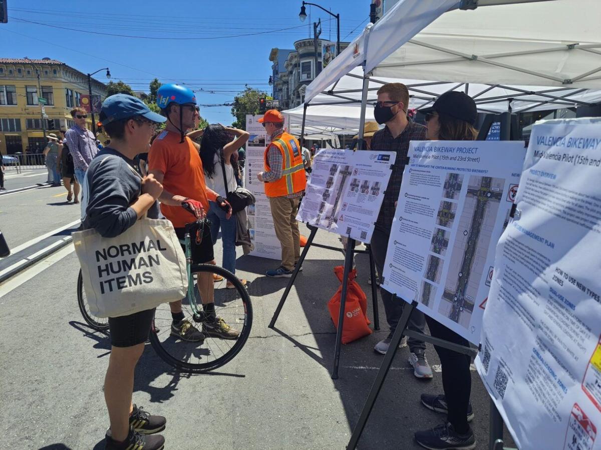 Outreach staff talk to community members in front of signs for the Valencia center-running bikeway Outreach staff talk to community members in front of signs for the Valencia center-running bikeway