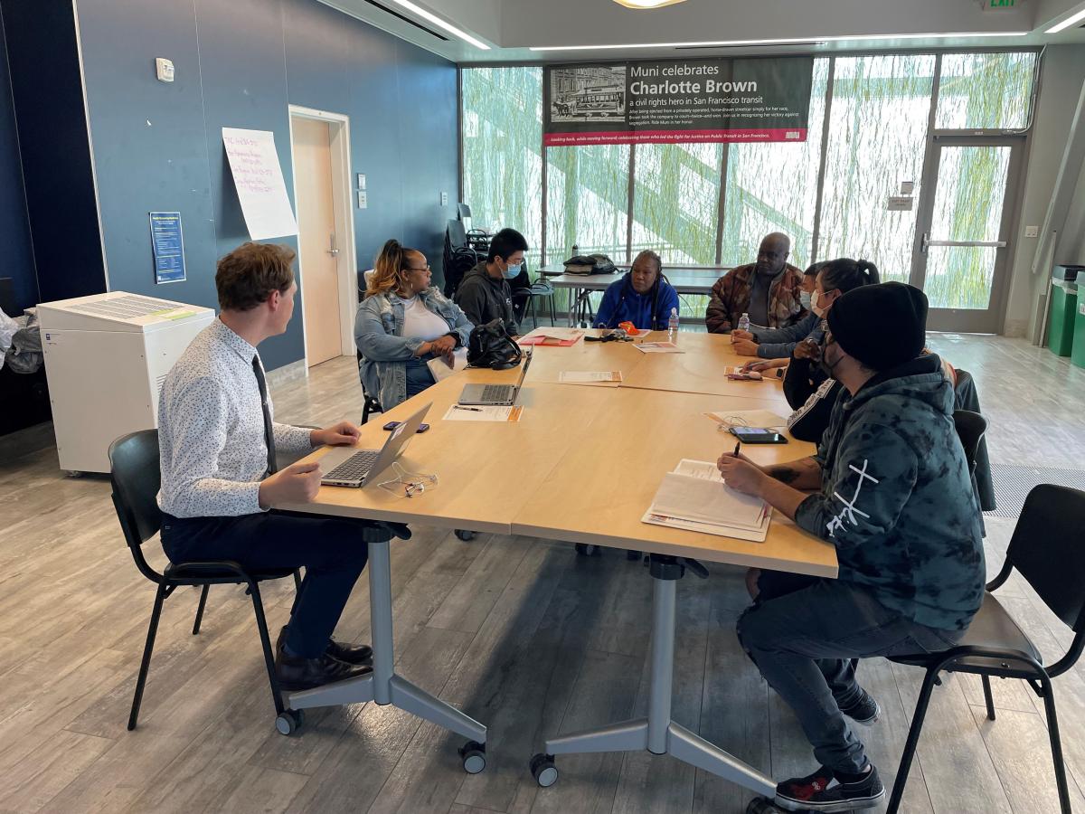 SFMTA staff and community partners sit at a table and chat in a conference room. SFMTA staff and community partners sit at a table and chat in a conference room.