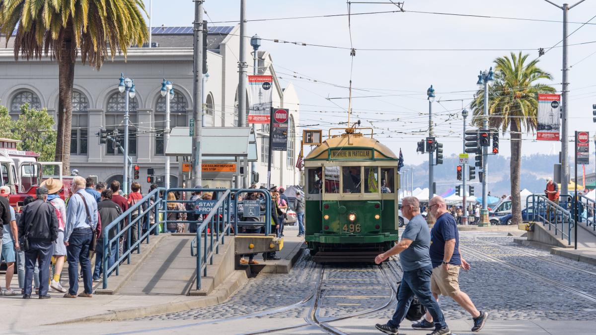 Green and cream historic streetcar stops near the Embarcadero. People cross an intersection in front of the streetcar. Green and cream historic streetcar stops near the Embarcadero. People cross an intersection in front of the streetcar.