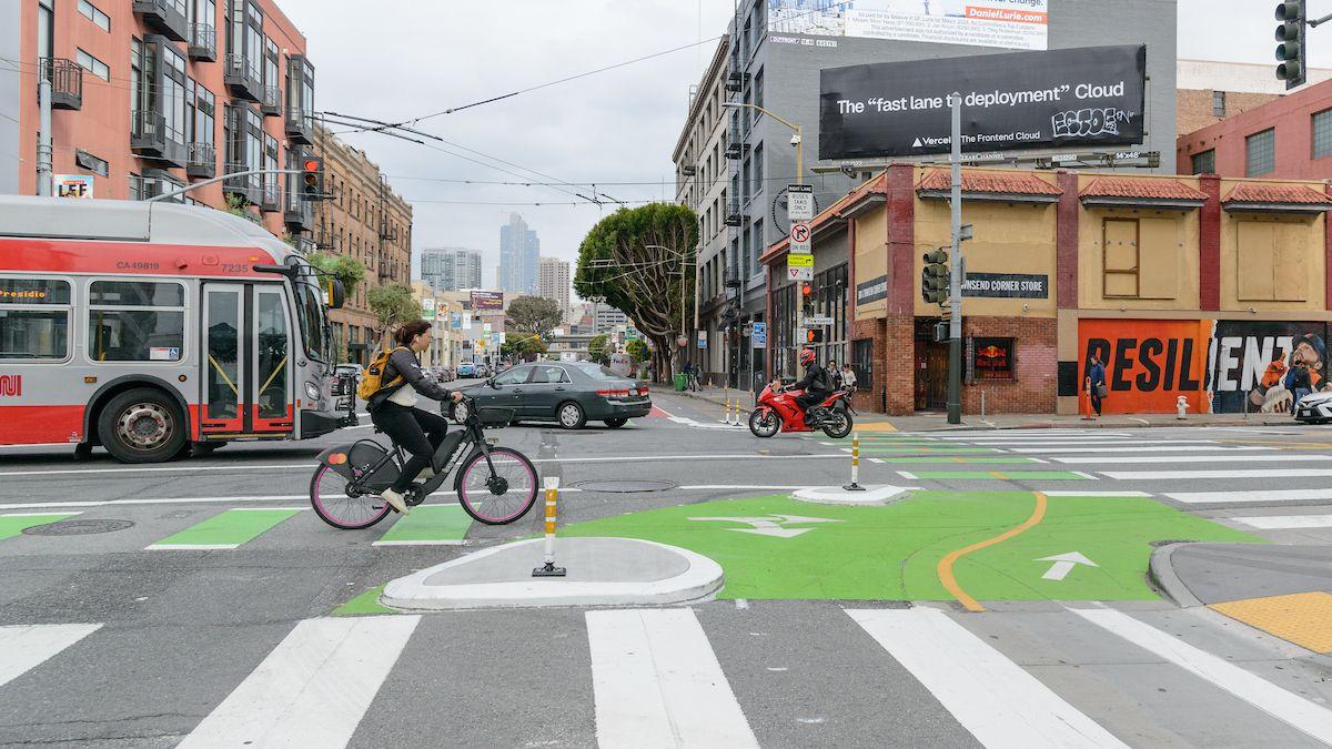 Person rides bike on a bikeway that' painted green in SoMa. In the background, a bus, vehicle and motorcycle cross the same intersection. Person rides bike on a bikeway that' painted green in SoMa. In the background, a bus, vehicle and motorcycle cross the same intersection.