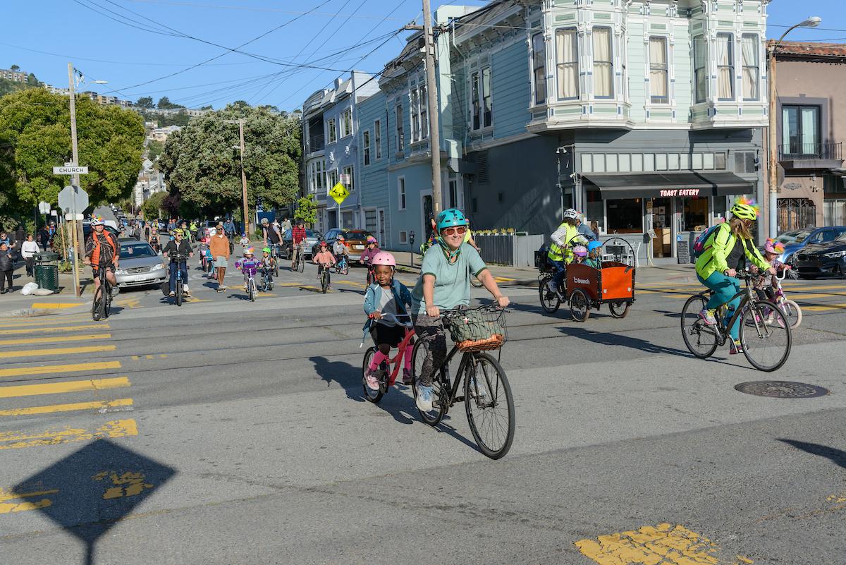 Adults and students ride their bikes, crossing a street on a sunny day. Adults and students ride their bikes, crossing a street on a sunny day.