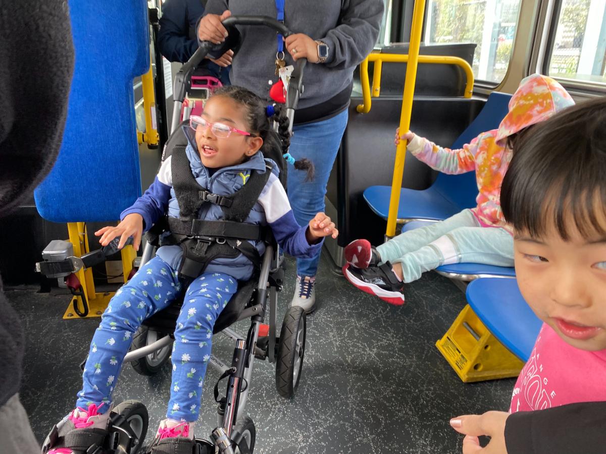 A passenger in a mobility stroller smiles near other young children on a bus. A passenger in a mobility stroller smiles near other young children on a bus.