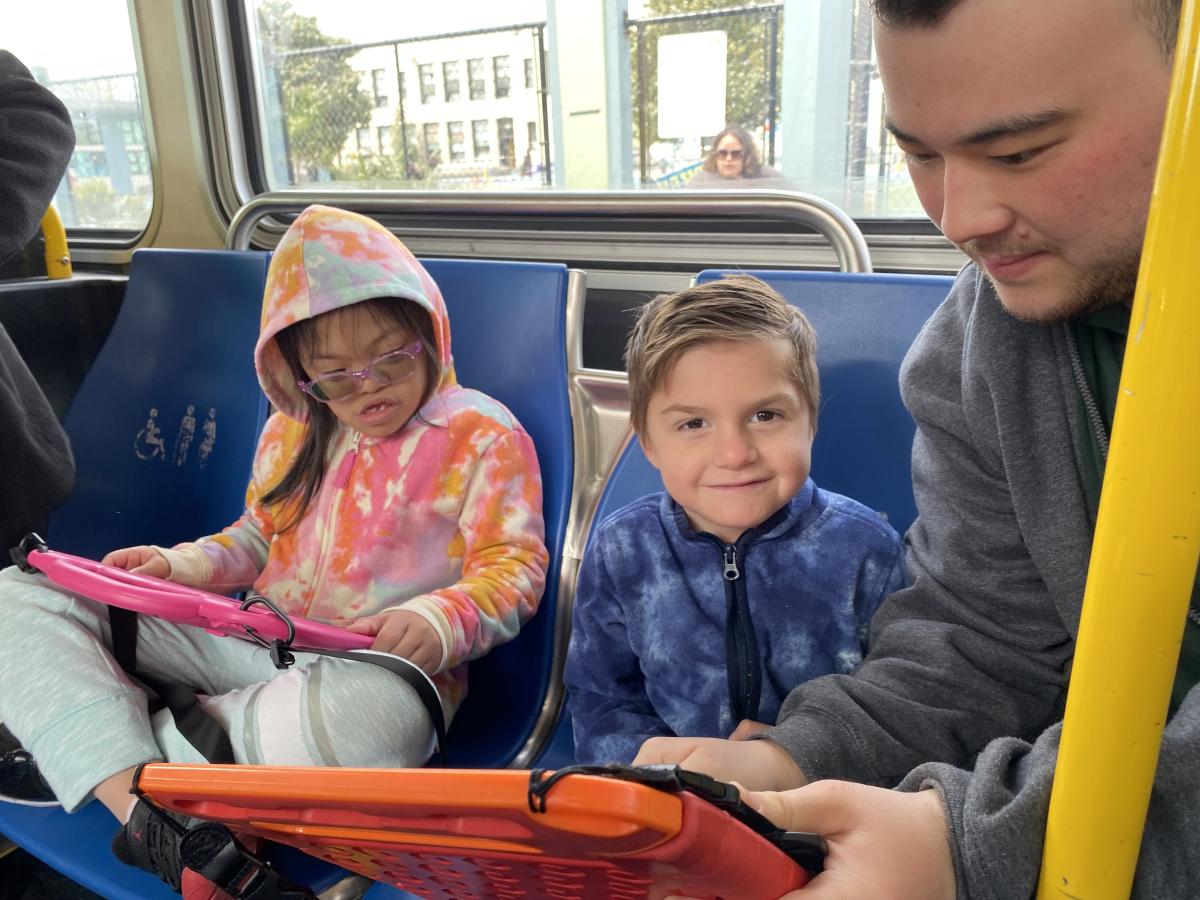 Two young children on a bus pay attention to an adult who shows them route information on a phone. Two young children on a bus pay attention to an adult who shows them route information on a phone.