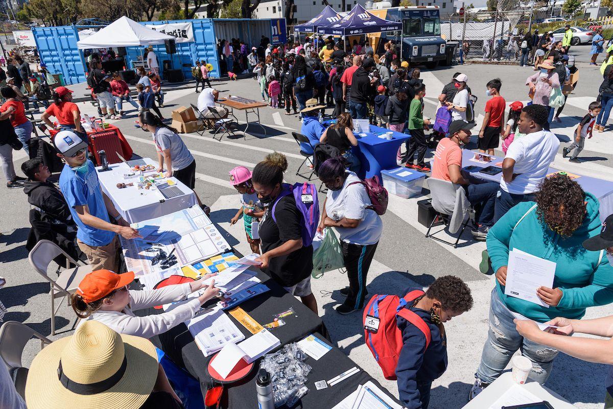 Overhead view of an outdoor public event where SFMTA staff and community members chat over tables about the Bayview Community Shuttle. Overhead view of an outdoor public event where SFMTA staff and community members chat over tables about the Bayview Community Shuttle.