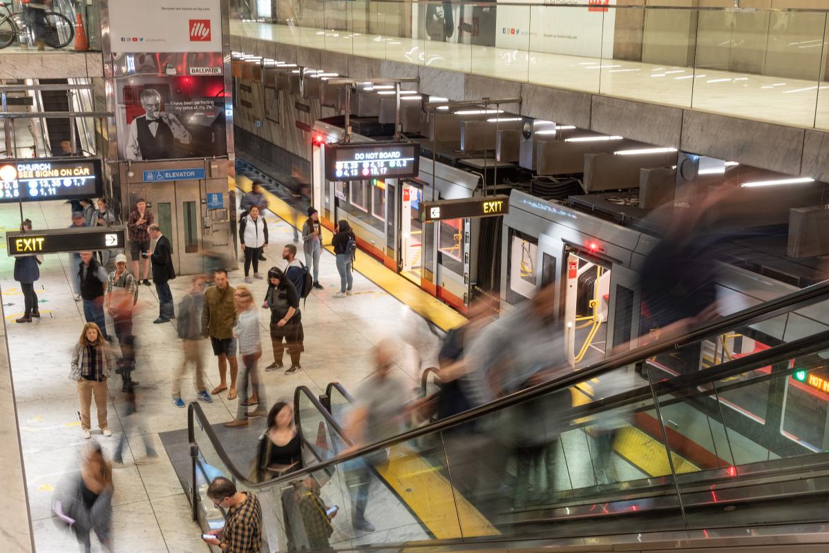 People wait for trains on a Muni platform as others exit on the escalator.