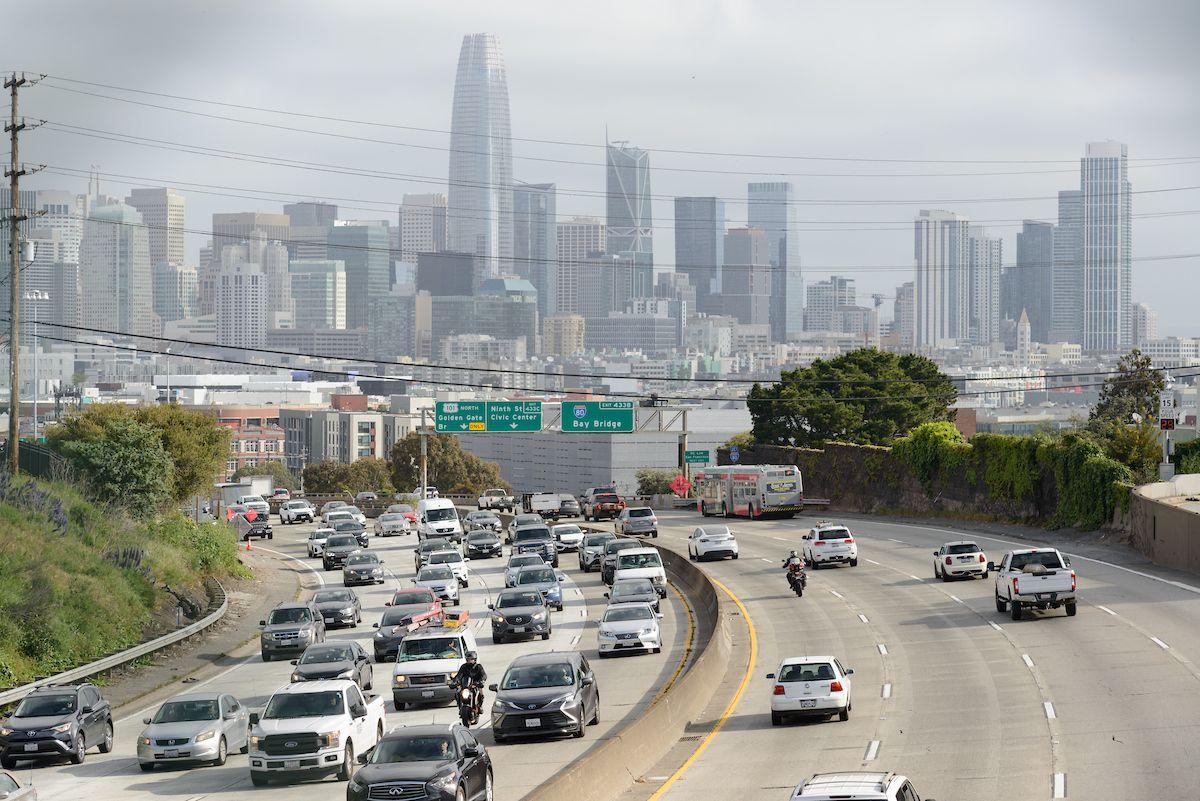 Cars drive on a highway with the downtown San Francisco skyline behind them. 