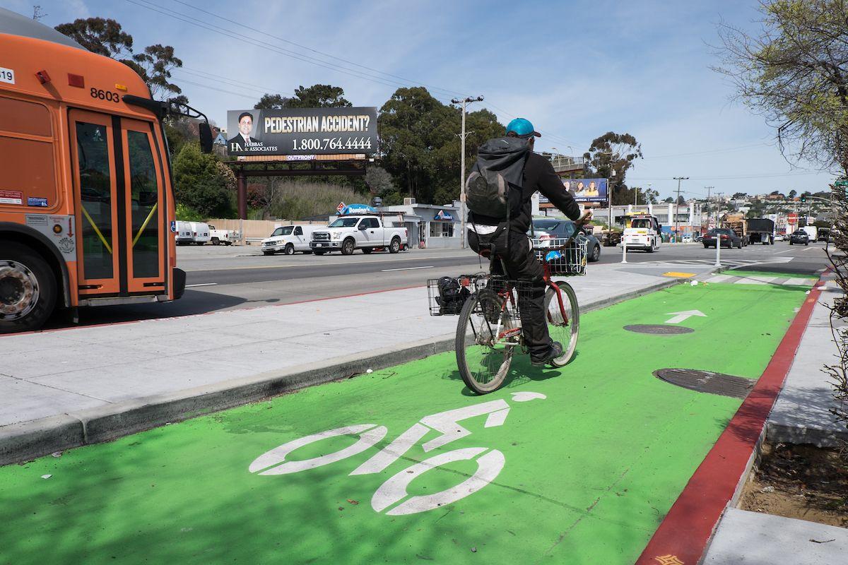 A photograph of a person riding a bicycle in a protected green bike lane on Bayshore Avenue. A bus can be seen on the other side of a boarding island.