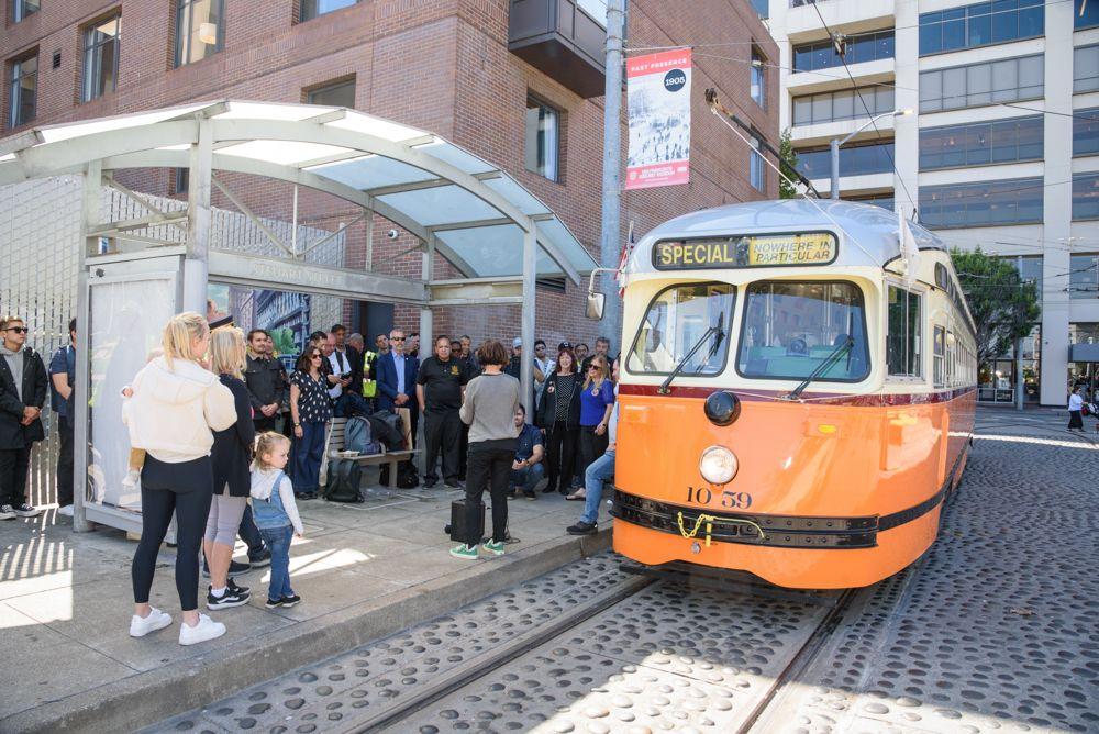 A large group of people is standing on a sidewalk, adjacent to a historic streetcar. The destination sign on the streetcar reads “nowhere in particular,” and it’s numbered 1059.
