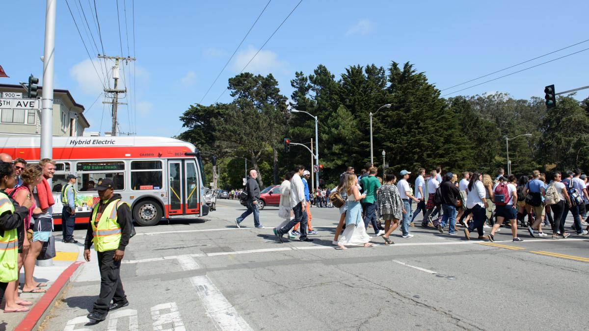 Many people cross an intersection toward Golden Gate Park during Outside Lands as a bus passes through the intersection nearby.