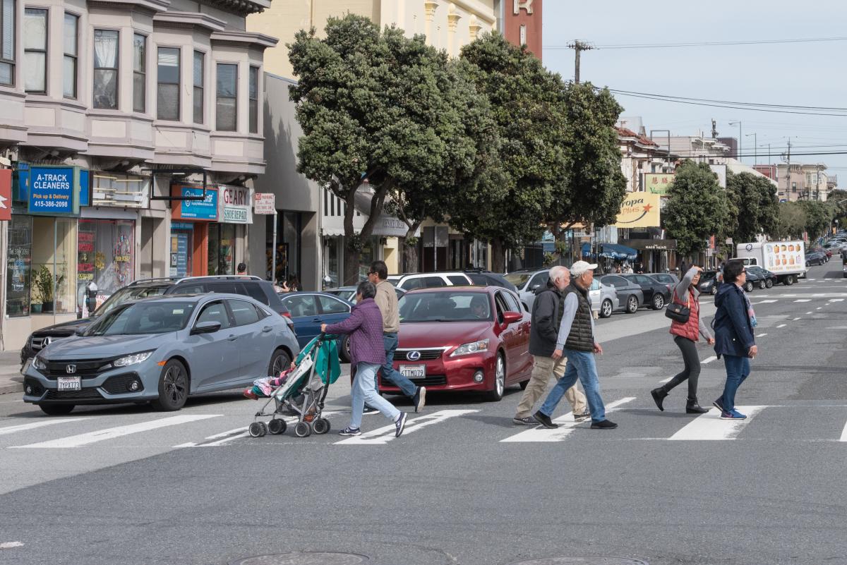 Several people cross Geary Boulevard as drivers behind the crosswalk. 