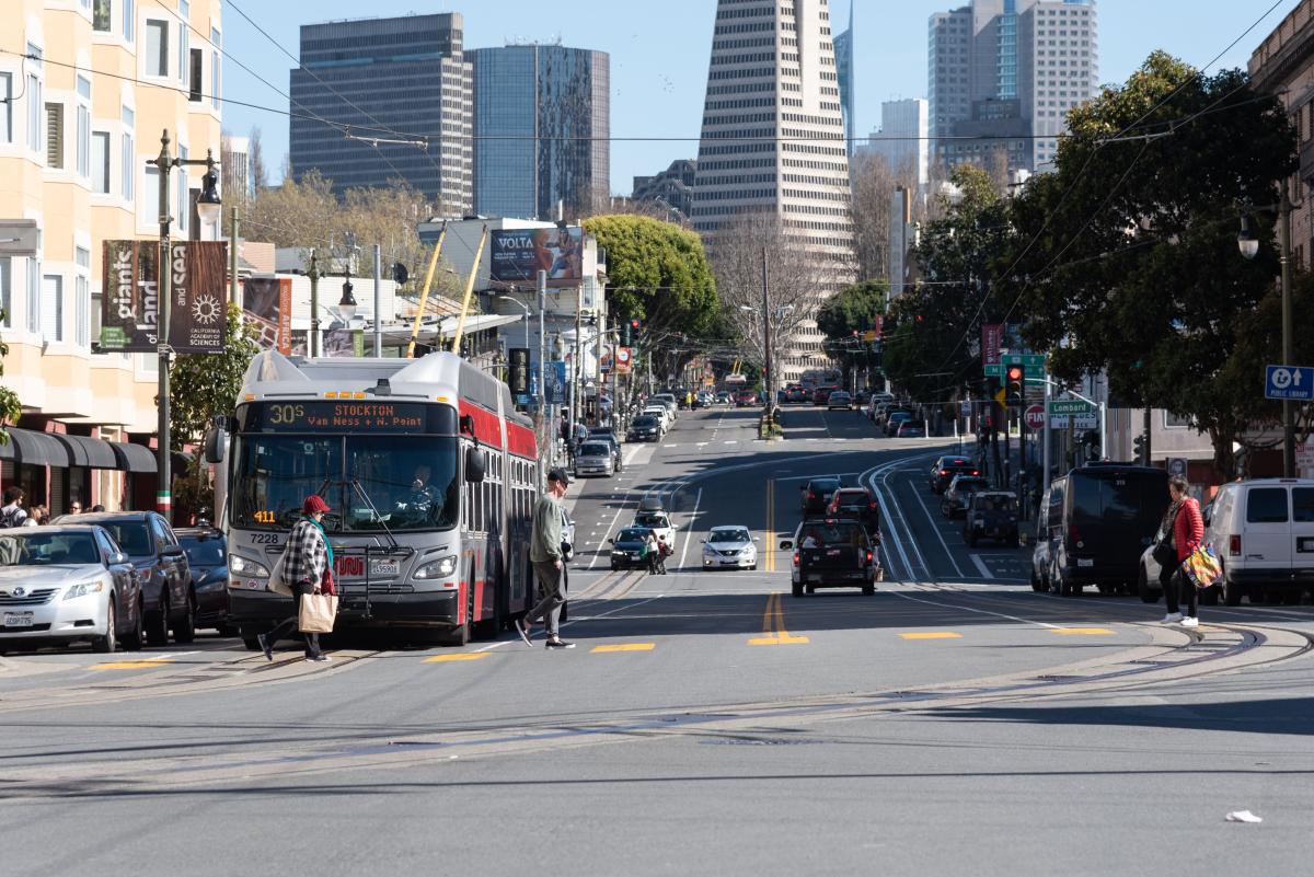 People cross Columbus Avenue as a bus waits at the intersection. 