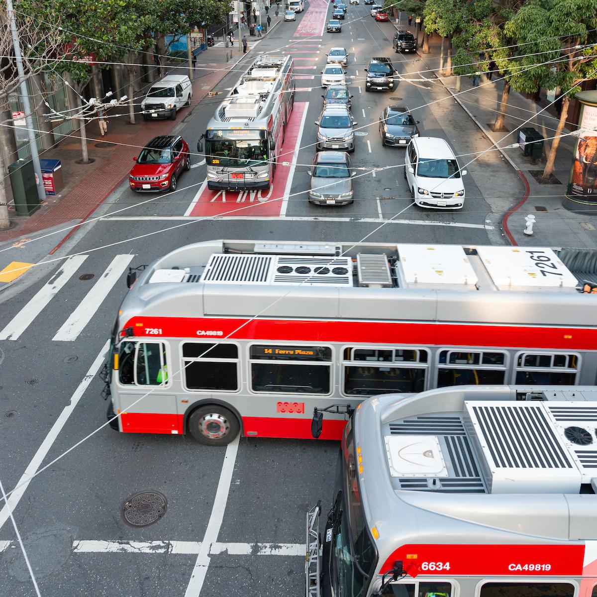 Two Muni buses drive through an intersection as a Muni bus and cars wait at the light.