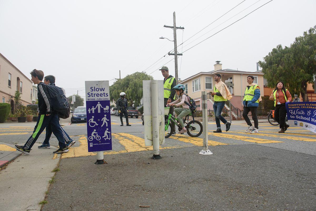 Students walk and ride bikes to cross a Slow Street alongside adults wearing yellow safety vests.