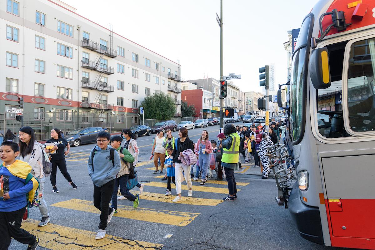 Students and families cross a street with help from a crossing guard. A bus pauses to let everyone through the intersecion. Students and families cross a street with help from a crossing guard. A bus pauses to let everyone through the intersecion.