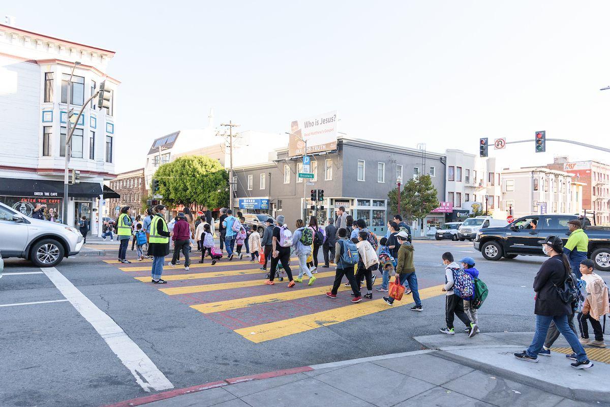 Students and families cross an intersection with help from crossing guards wearing yellow safety vests. Students and families cross an intersection with help from crossing guards wearing yellow safety vests.