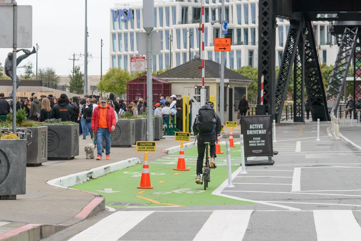 Person rides a bike on a bikeway that's painted green. People walk on the sidewalk nearby.