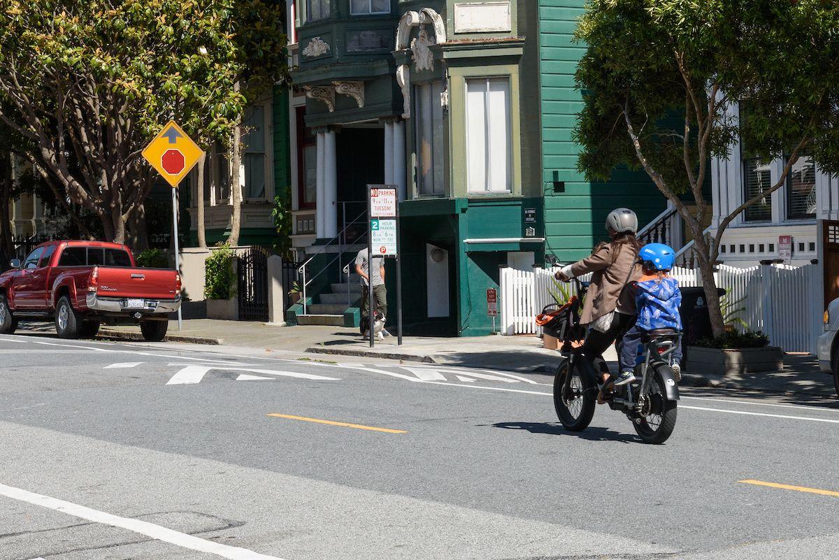 A woman and young child ride an electric bike down a city street with colorful homes in the background.