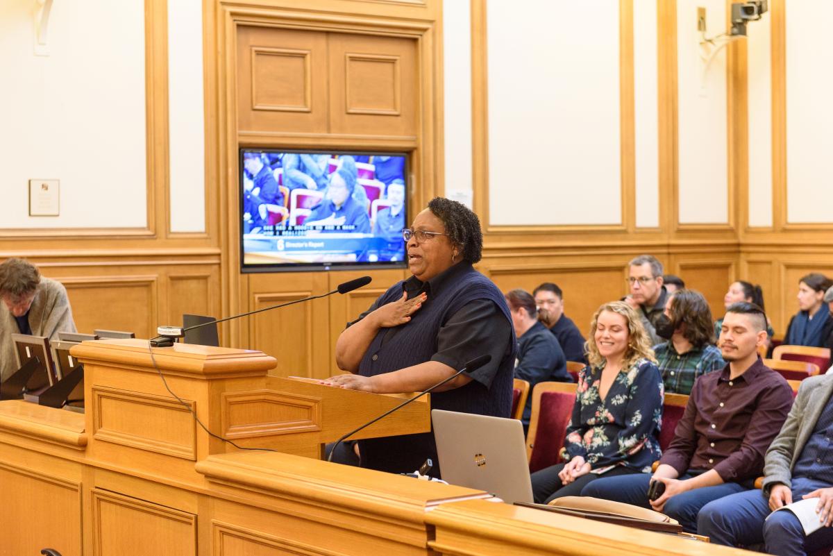 Denise Green holds speaks at a mic for the SFMTA Board of Directors special recognition award. A crowd sits in rows behind her.