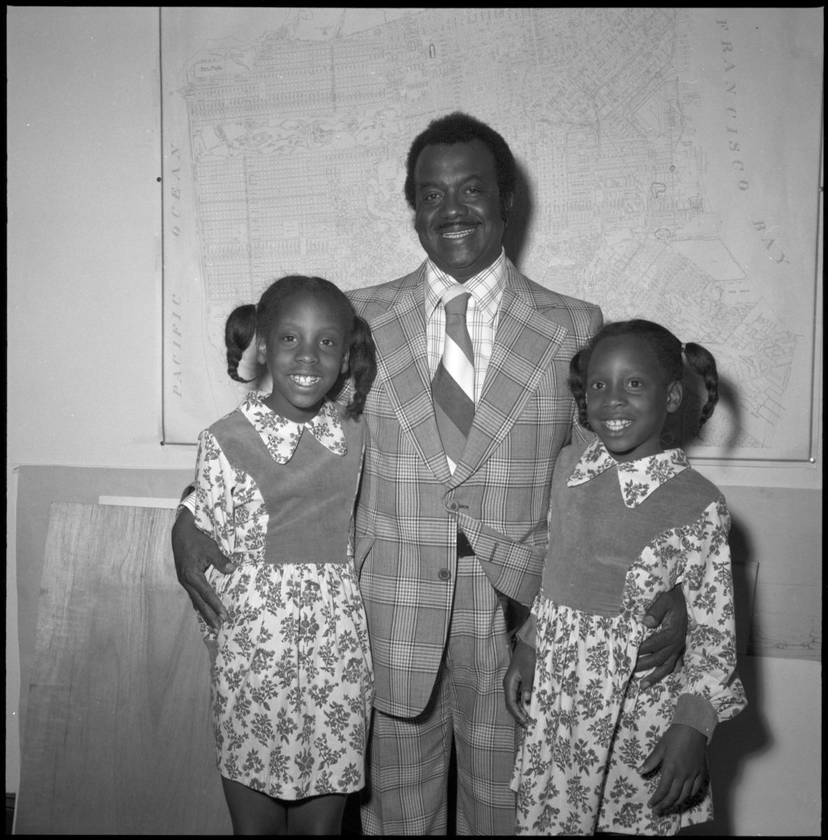 Childhood photo of Denise Green and her sister with their grandfather, Curtis Green. The three smile as they stand against a wall that has a map of San Francisco.