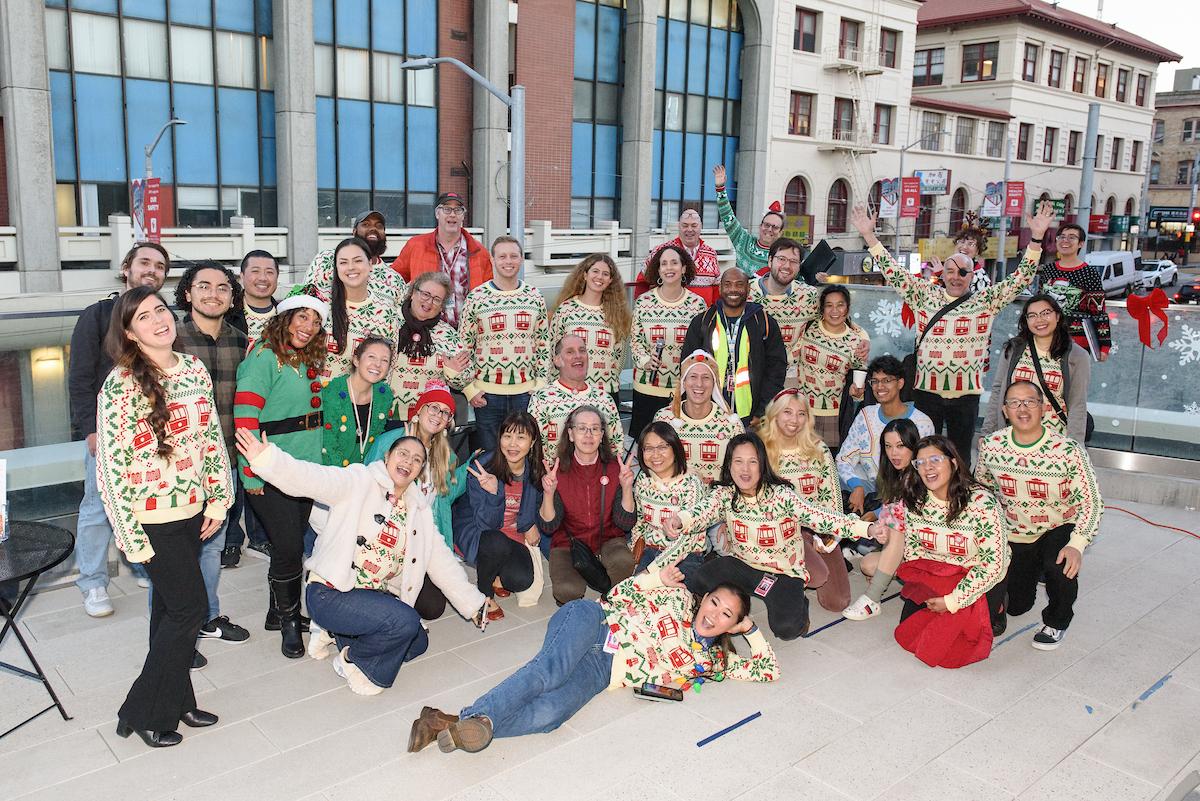 Several people wearing the Muni Icons of San Francisco sweater pose in front of buildings.
