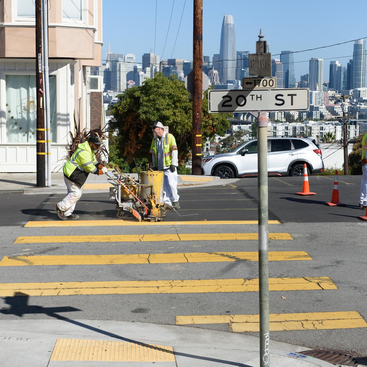 SFMTA crews complete road work on a street overlooking downtown buildings.
