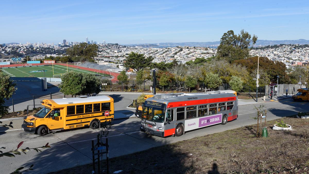 A Muni bus and a school bus use the same street overlooking a school's football field as well as downtown San Francisco.