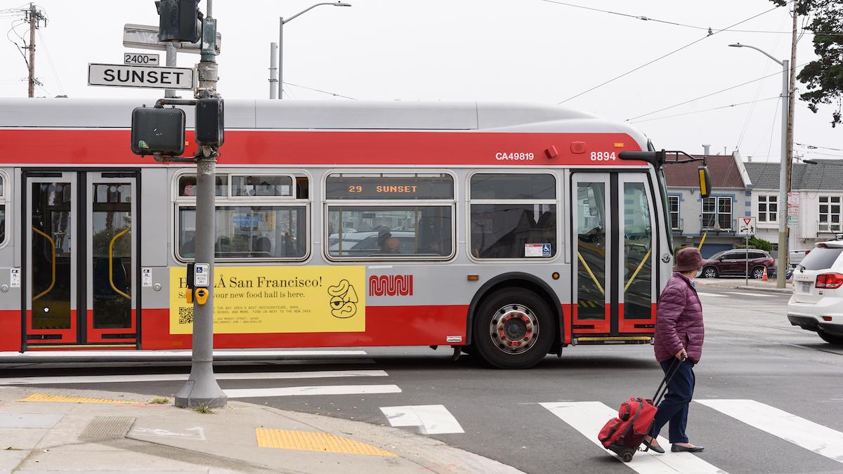 29 Sunset drives through an intersection. Nearby, a woman crosses the street.