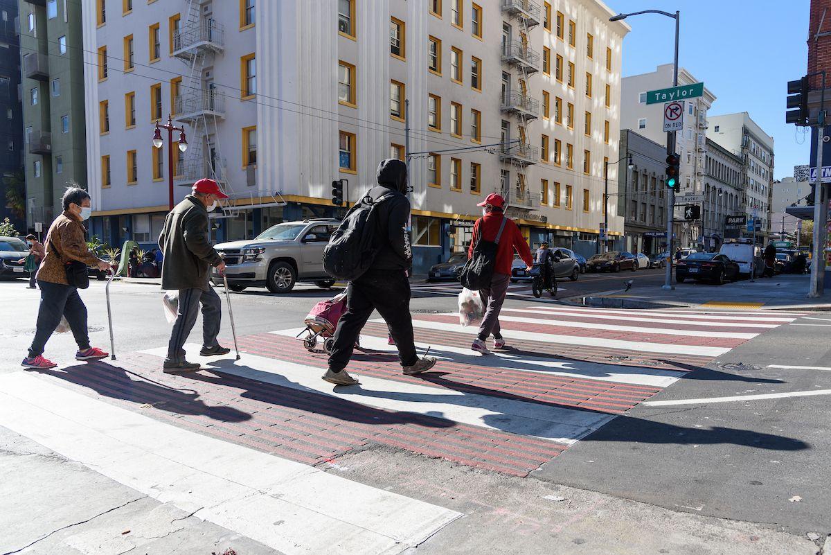 Four people cross a downtown sidewalk painted red and white as cars pass by in the background.