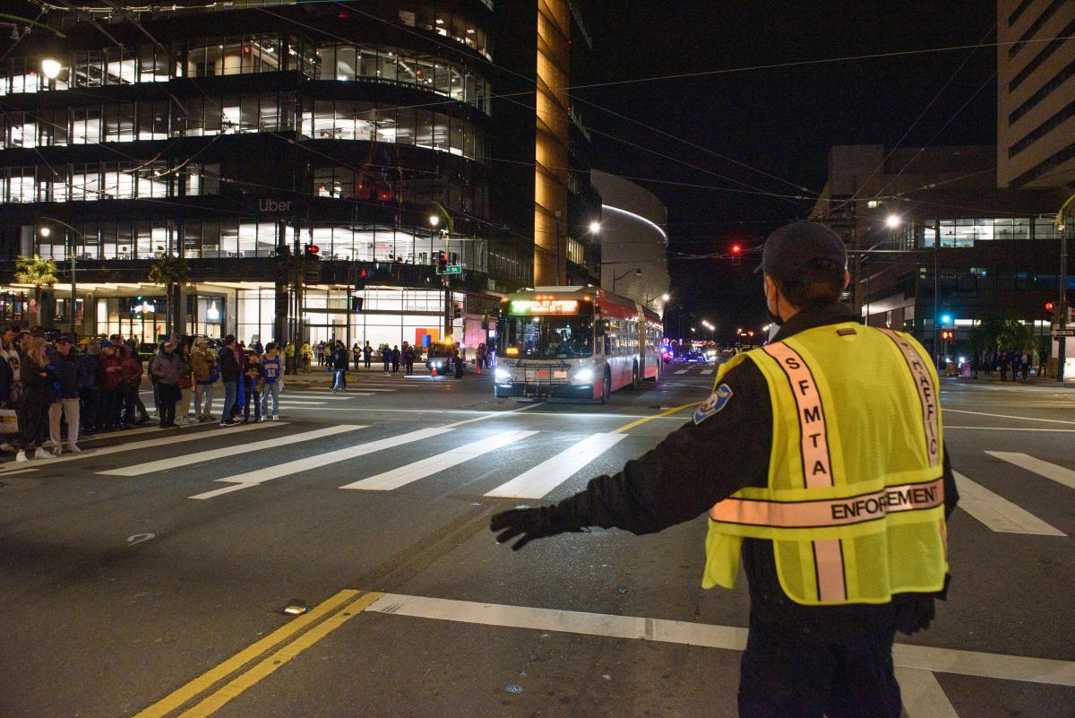 Parking control office wearing a yellow SFMTA vest helps direct traffic as a crowd waits on a corner and a Muni bus approaches.