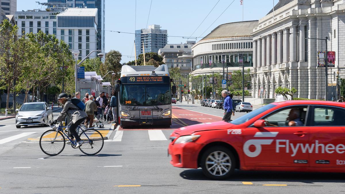 Closeup of an intersection with a person biking, person driving a red cab and bus stopped to pick up passengers.