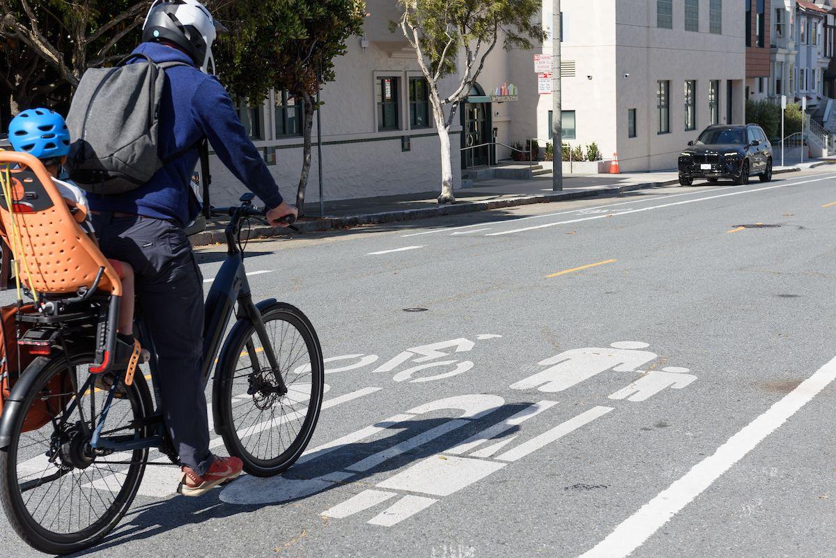 Adult rides a bike with a small child in the back seat on a shared roadway.