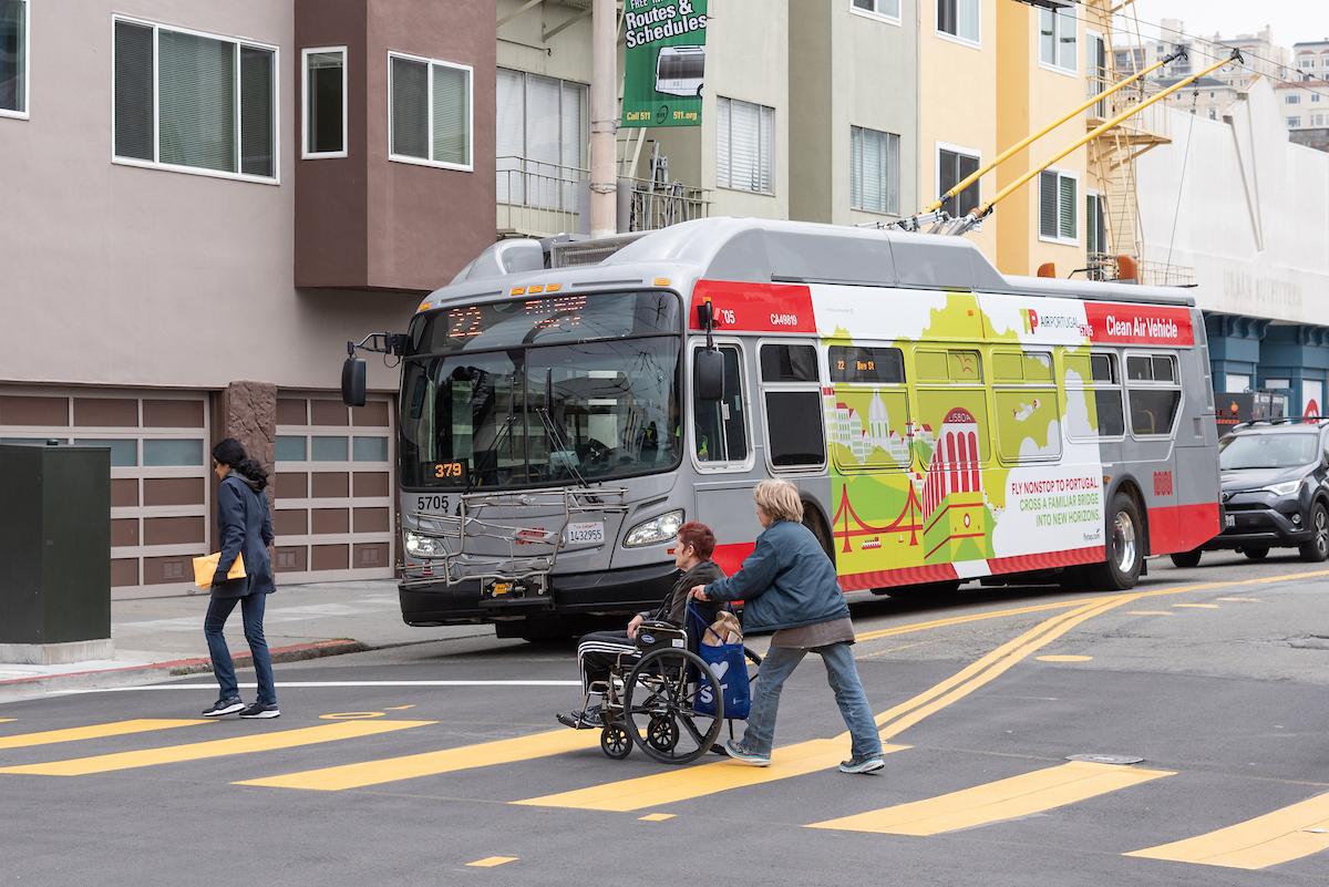 Closeup of a crosswalk as a person pushes another person in a wheelchair and another person walks.